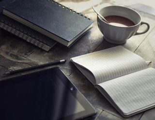 On the table is an open notebook, a cup of coffee, a tablet, a pen and two books on top of each other
